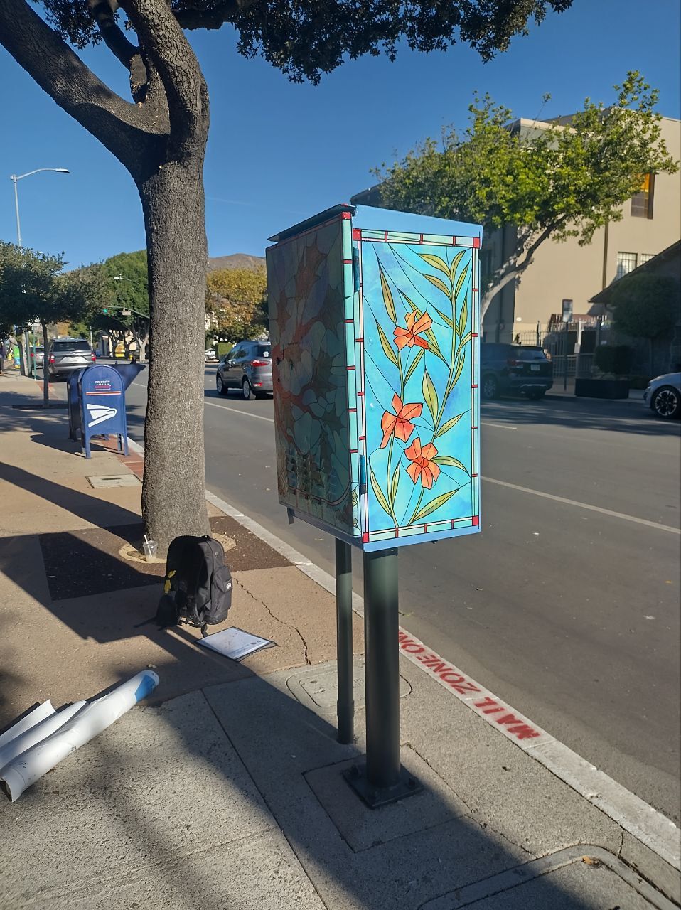 Painted utility box on a sunny city street beside a tree and parked cars