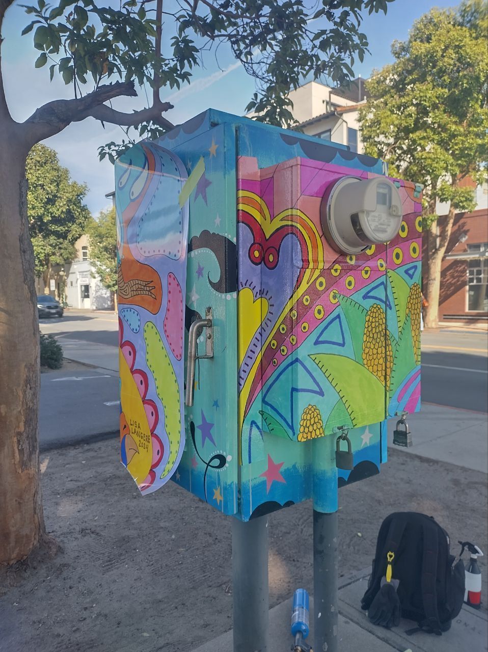 Colorful graffiti-painted utility box on a sidewalk under trees, with a backpack beside it.