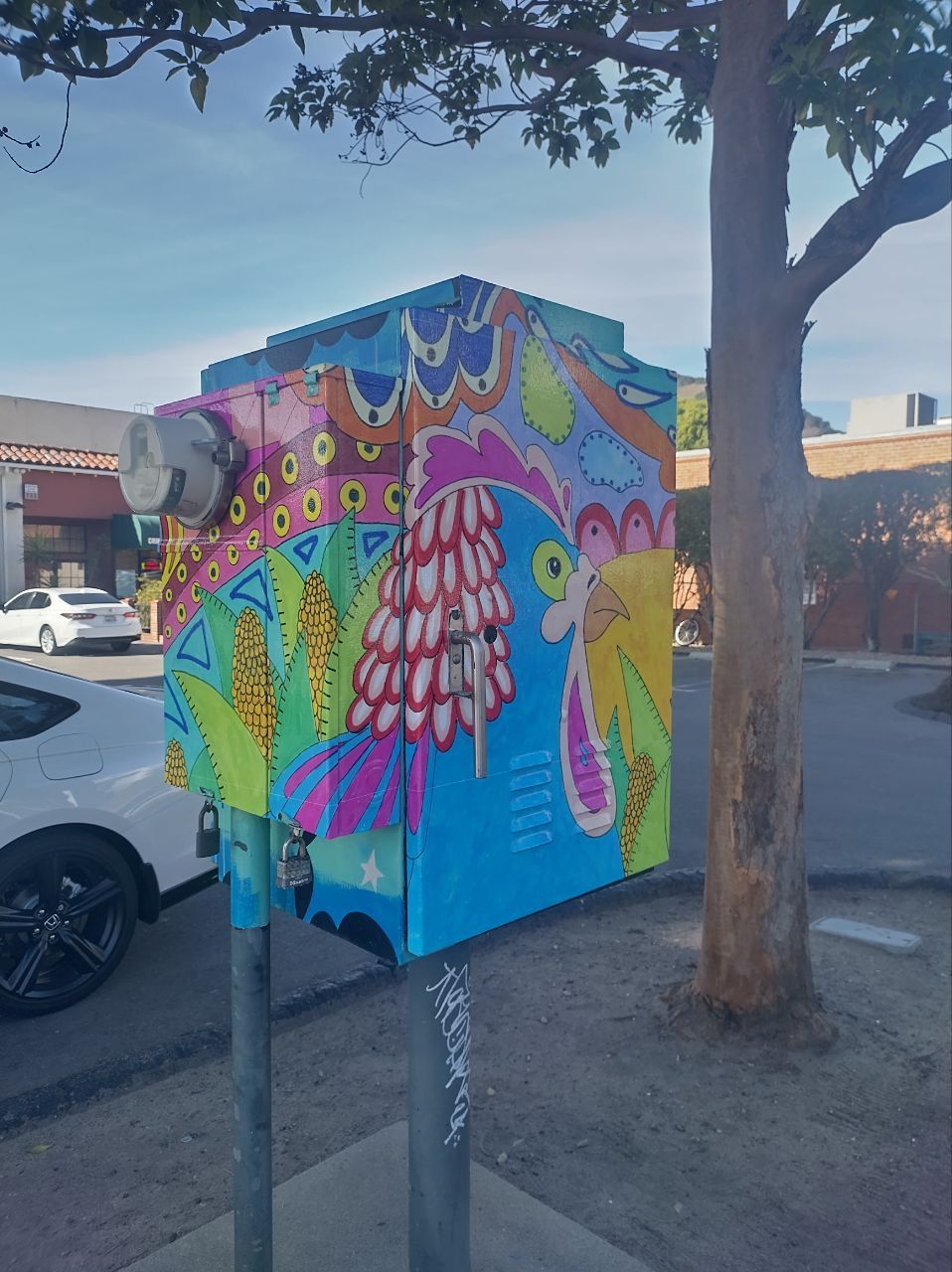 Colorful owl mural on a utility box beside a sidewalk and tree, with parked cars nearby.