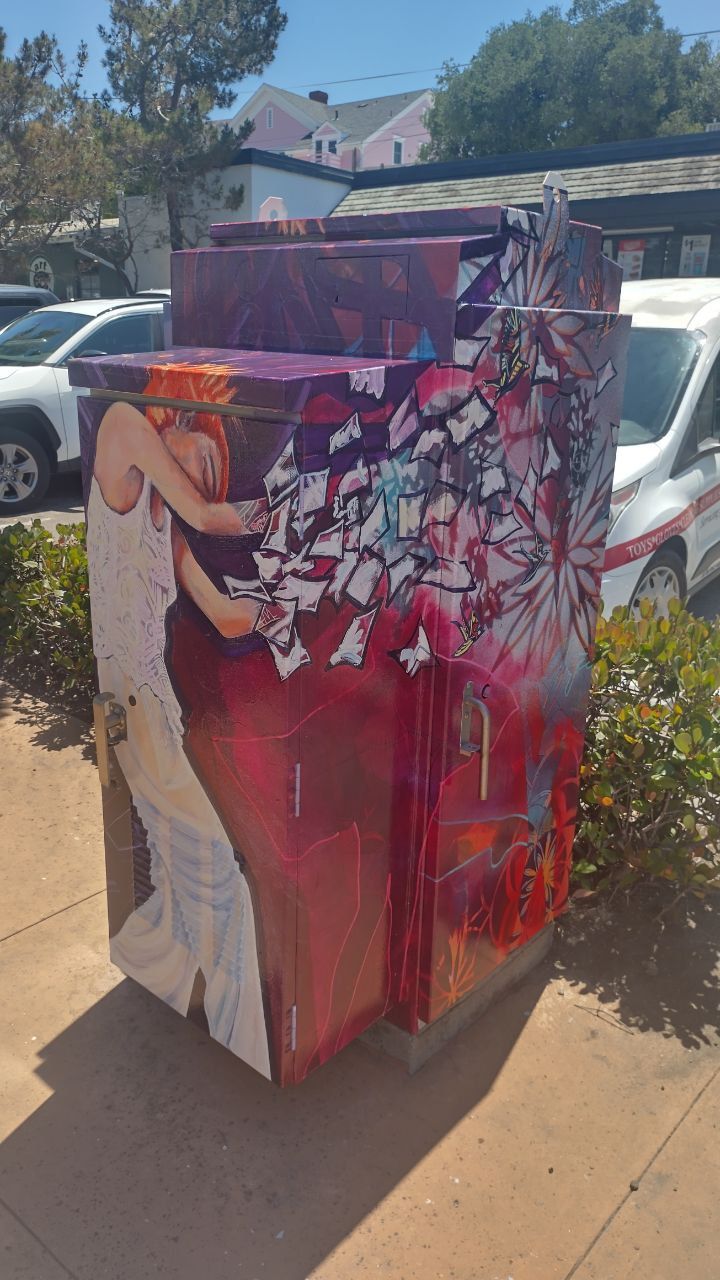 Painted utility box with red and white abstract mural on a sunny sidewalk near parked cars