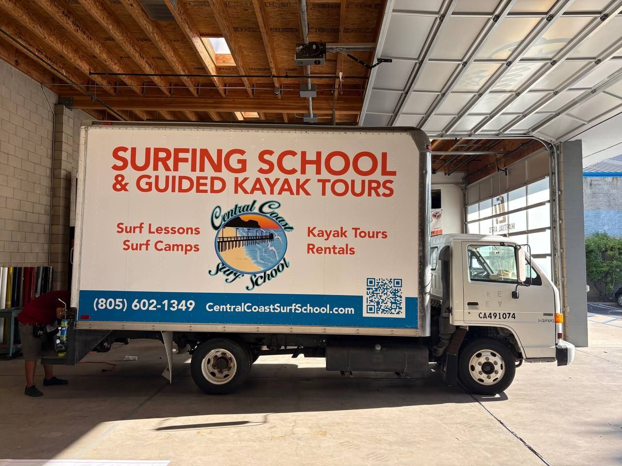 White truck with “Surfing School & Guided Kayak Tours” signage parked in a covered area