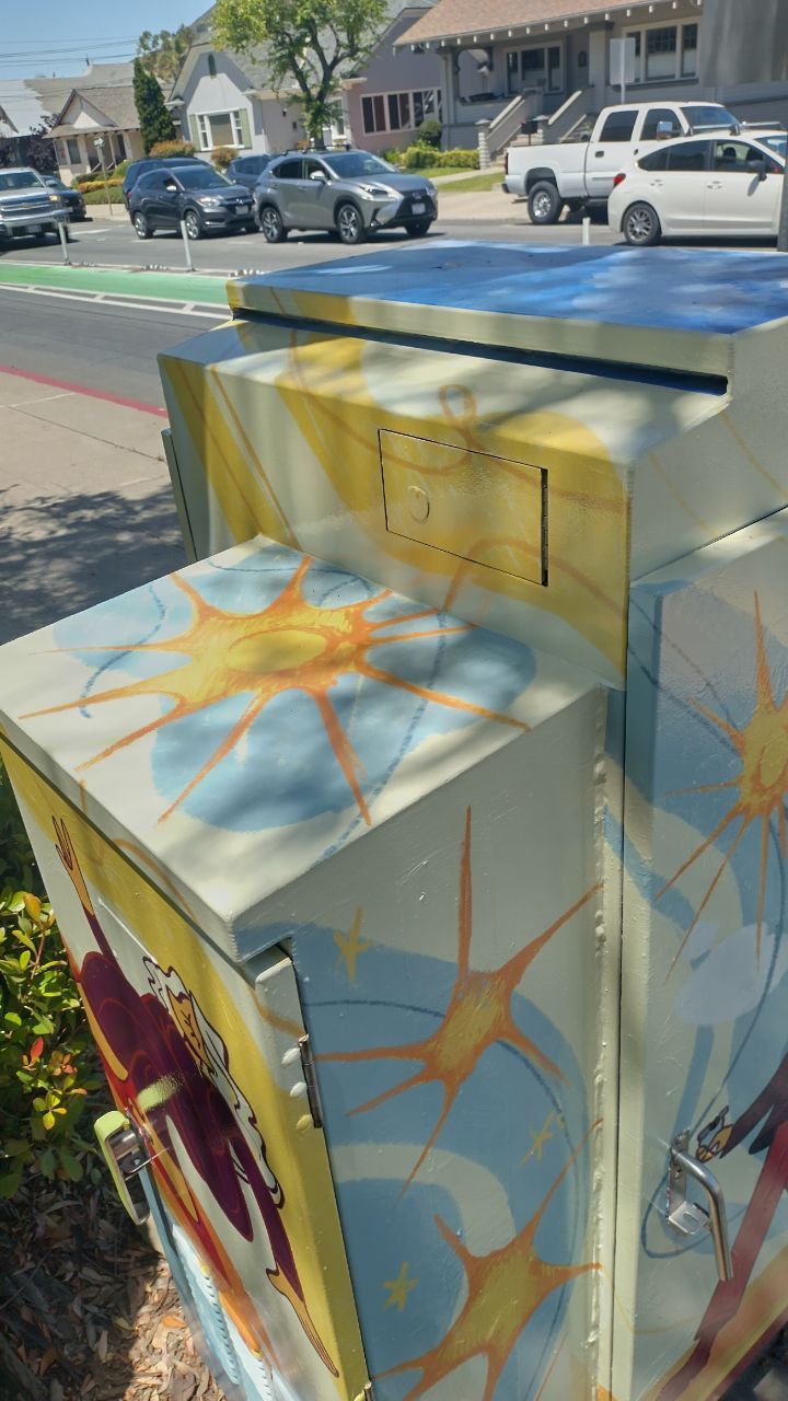Painted utility box on a sunny sidewalk with blue, yellow, and sunburst designs