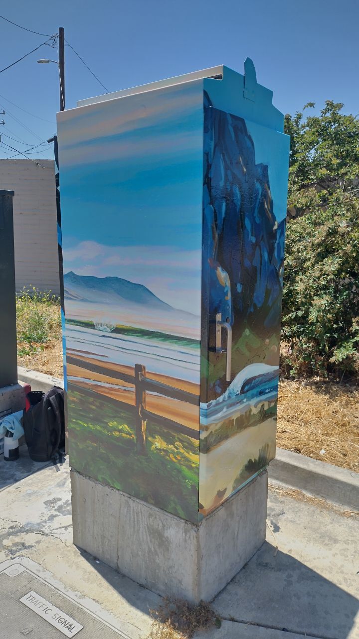 Utility box painted with a coastal landscape mural of ocean, mountains, and a fence.