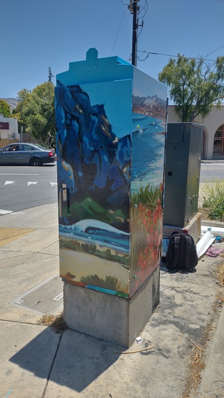 Painted utility box with colorful mountain and landscape mural on a sunny sidewalk near parked cars