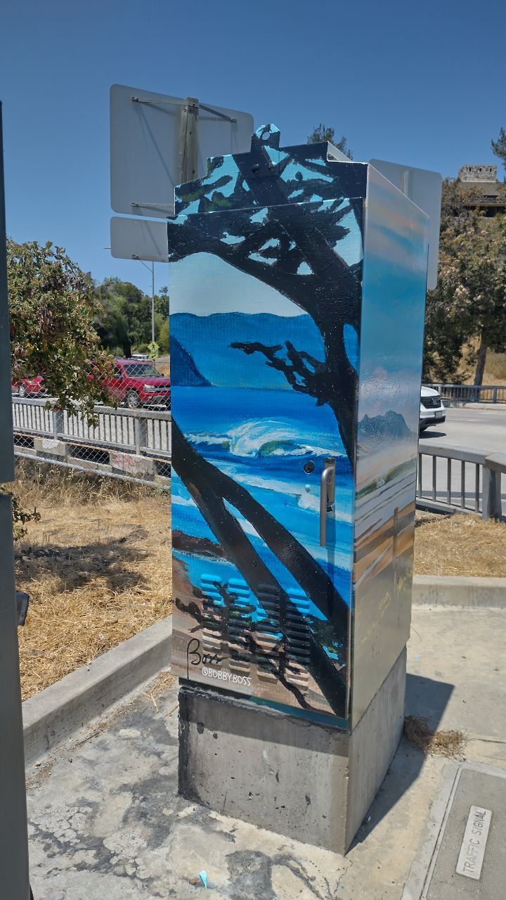 Painted utility box with blue ocean and tree silhouette mural on a sunny roadside curve