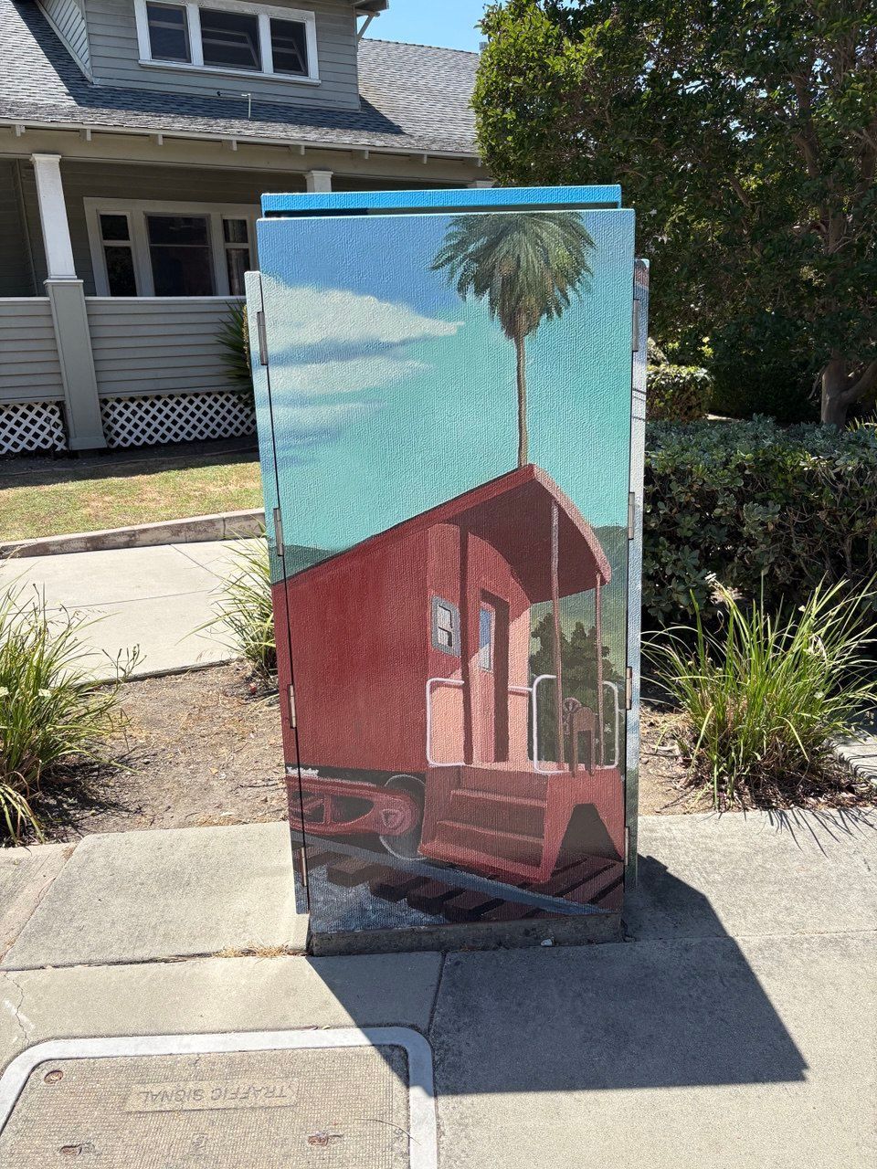 Mural-painted utility box with a red house and palm tree on a sidewalk in front of a home