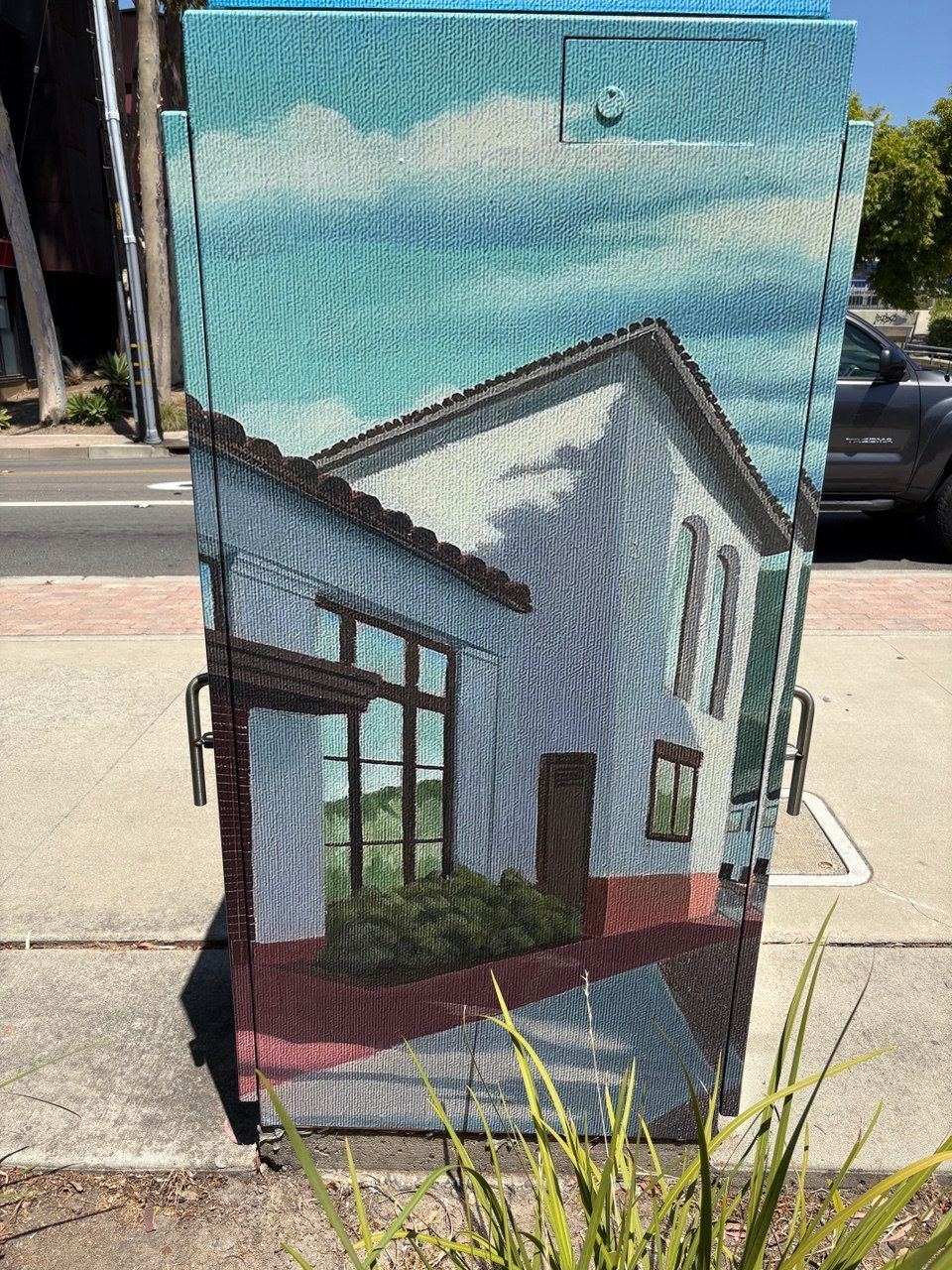 Painted utility box with a white house mural beside a sidewalk and street