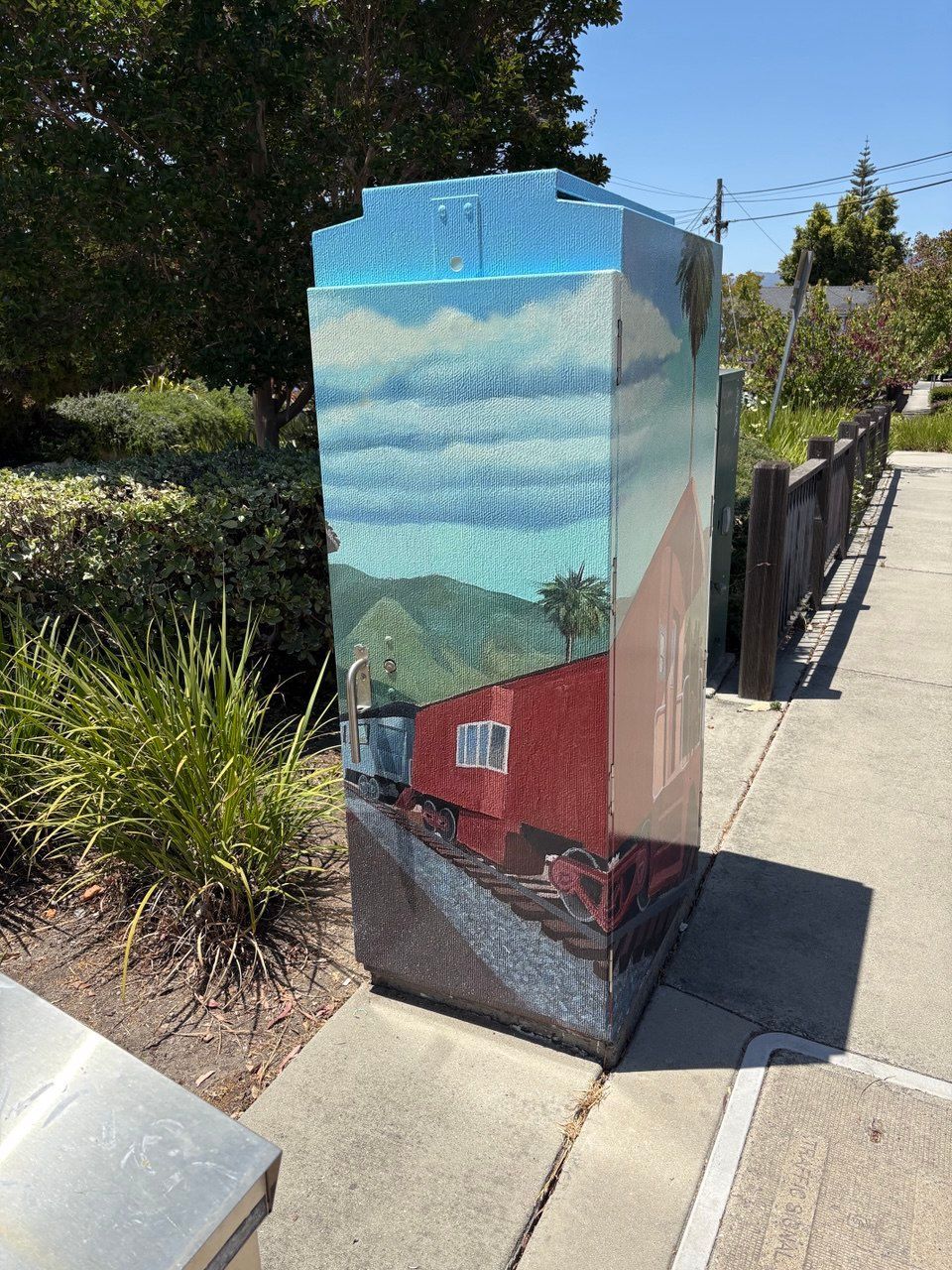 Painted utility box with a seaside mural beside a sidewalk and fence on a sunny day