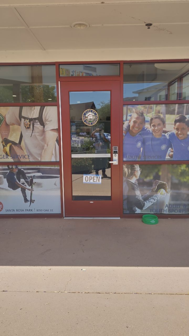 Storefront with red-framed glass doors and sports-themed window murals in a shopping center.