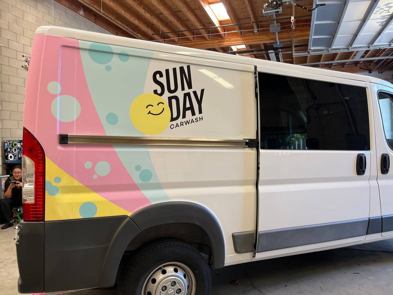 White van with colorful “Sun Day” mural and open side door at an indoor event.
