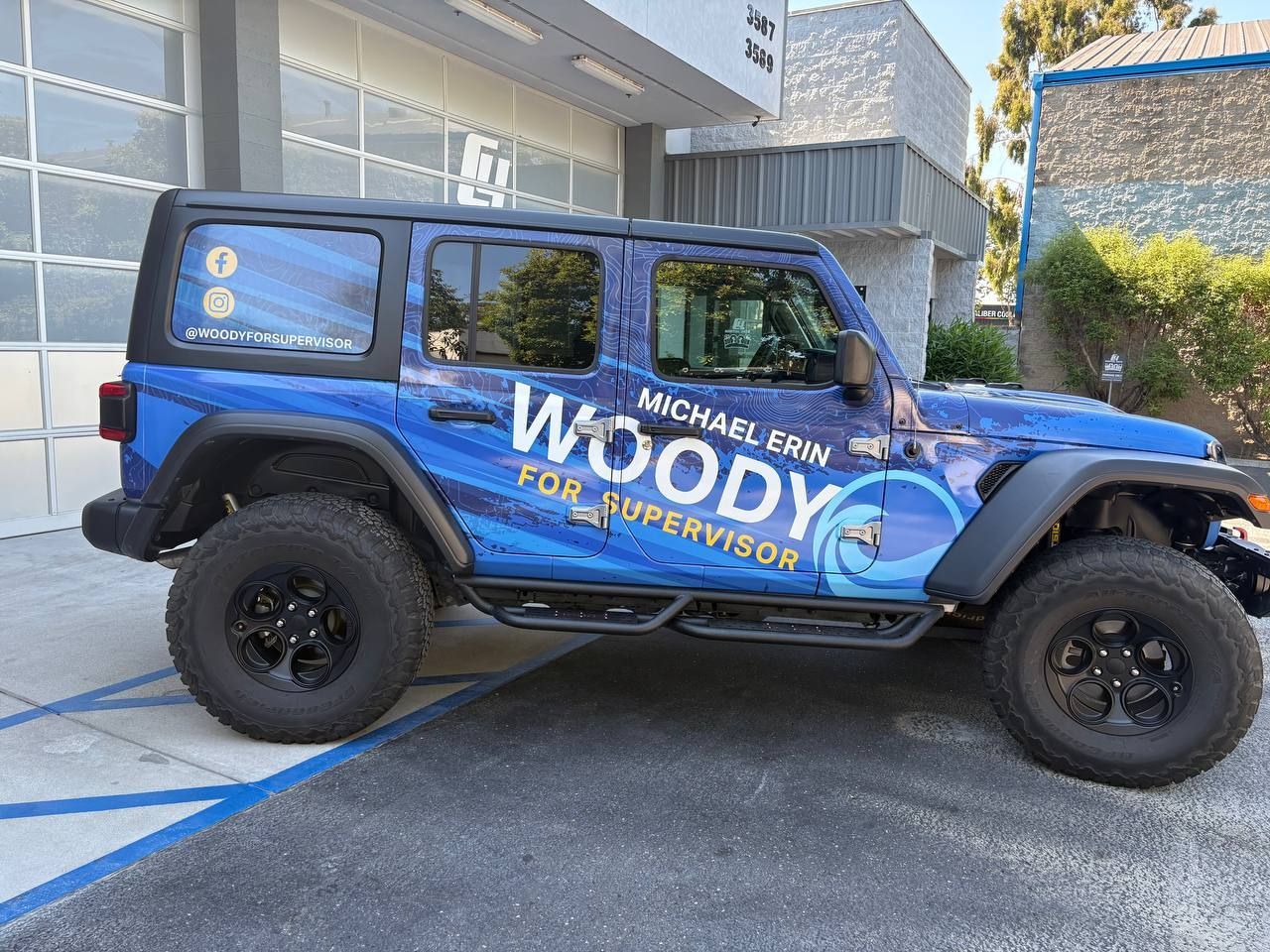 Blue Jeep Wrangler wrapped in Woody Woodpecker graphics parked on a street by a modern building.