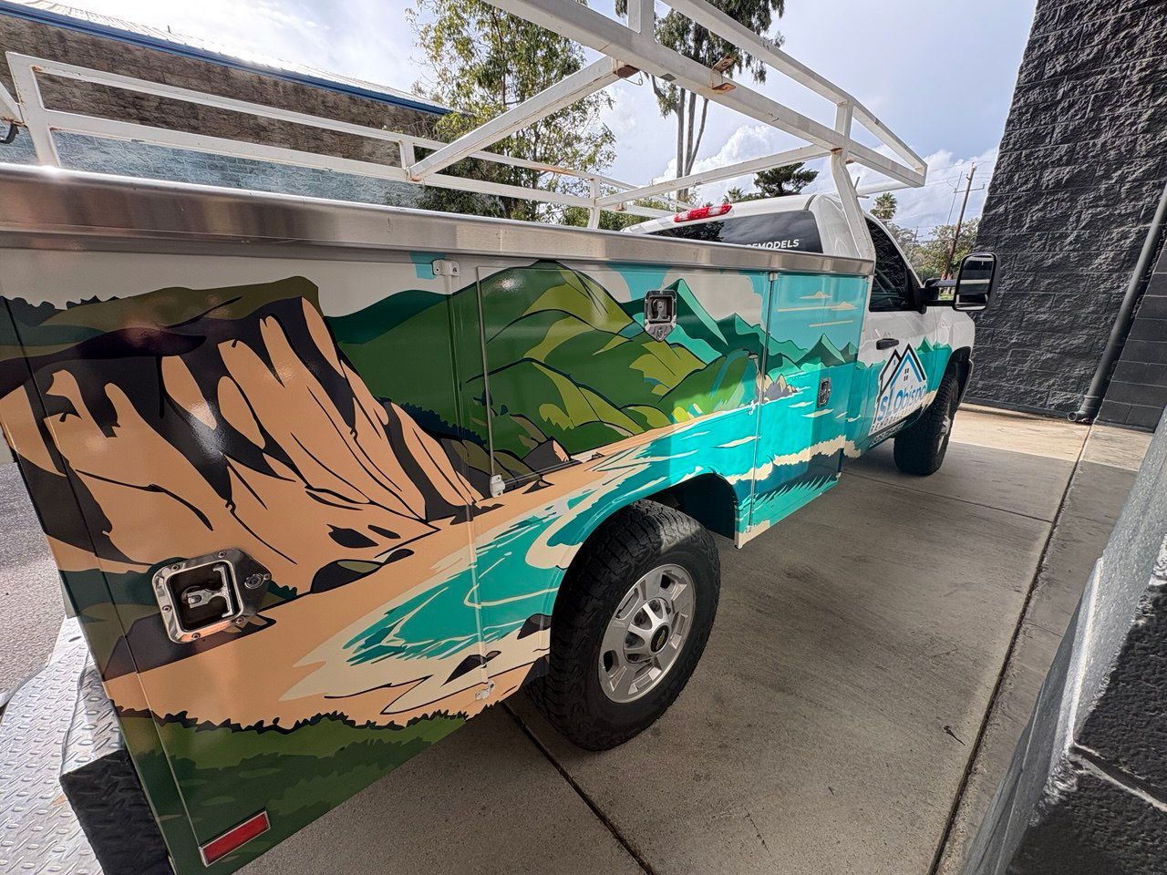 Colorful mural on the side of a truck parked in a driveway, with mountains, water, and a car wheel visible