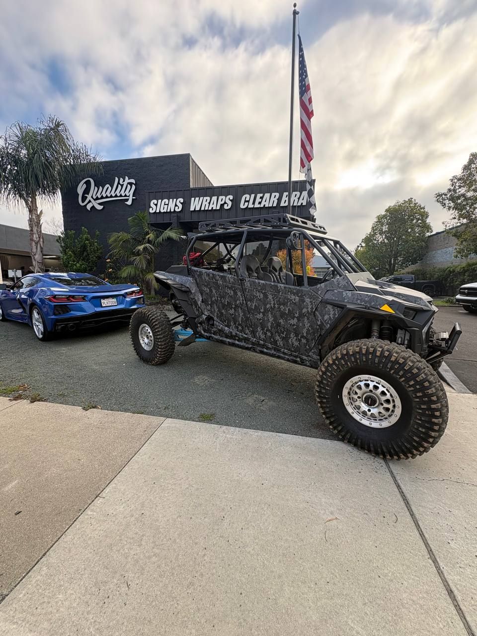 Camouflage off-road buggy with large tires parked beside a blue sports car at an outdoor event