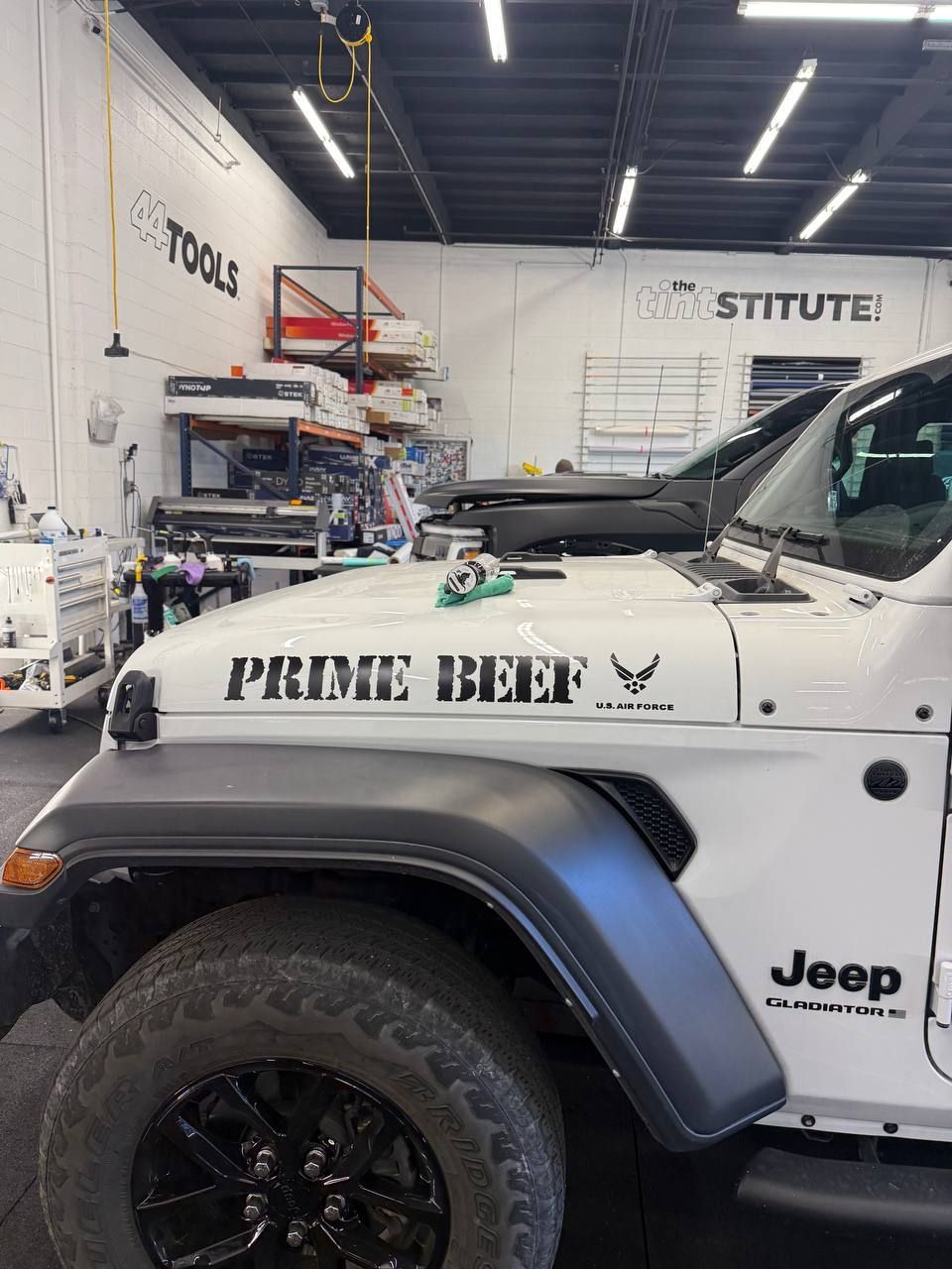 White Jeep with “PRIME BEEF” text in a garage shop, showing the front fender and tire.