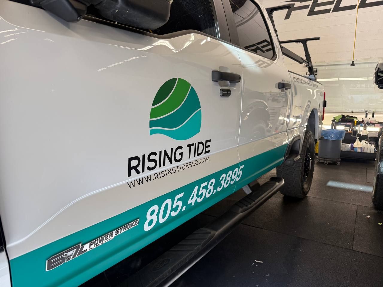White boat with Rising Tide logo and teal phone-number stripe inside a boat showroom.