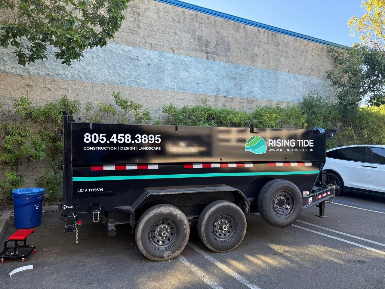Black utility trailer with landscaping company branding parked in a lot beside a building