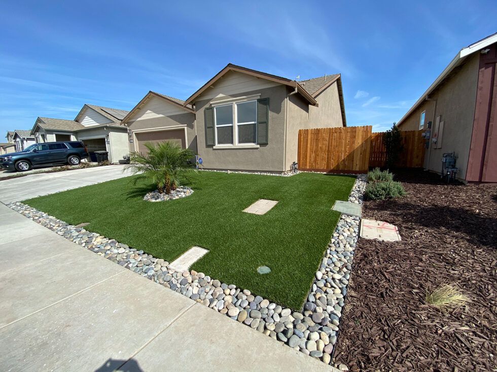 Suburban front yard with artificial turf, stone edging, and a small desert plant beside a house driveway