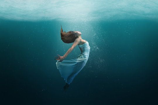 Woman in a blue dress floating underwater, curled in a teal ocean scene