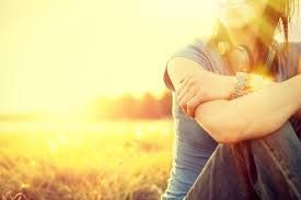 Person sits outside, arms crossed, bathed in sunlight. Headphones, field in background.