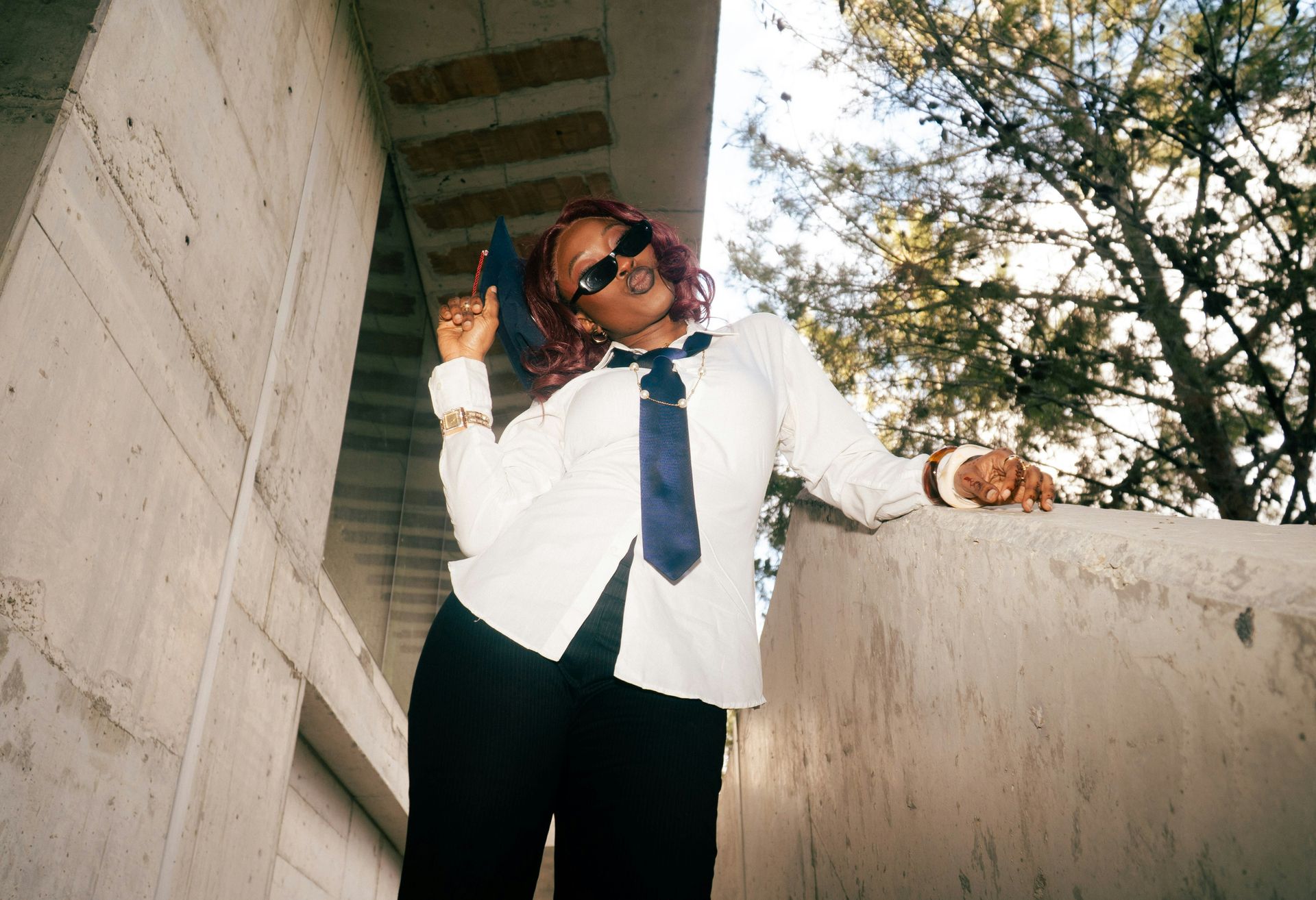 Person in white shirt and black pants posing under a concrete overpass, wearing sunglasses and a blue tie.