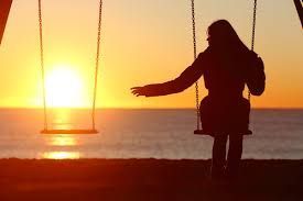 Silhouette of person on swing set facing the ocean at sunset; empty swing beside them.