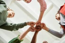 Hands of several people stacked together in a circle, viewed from below against a bright ceiling.