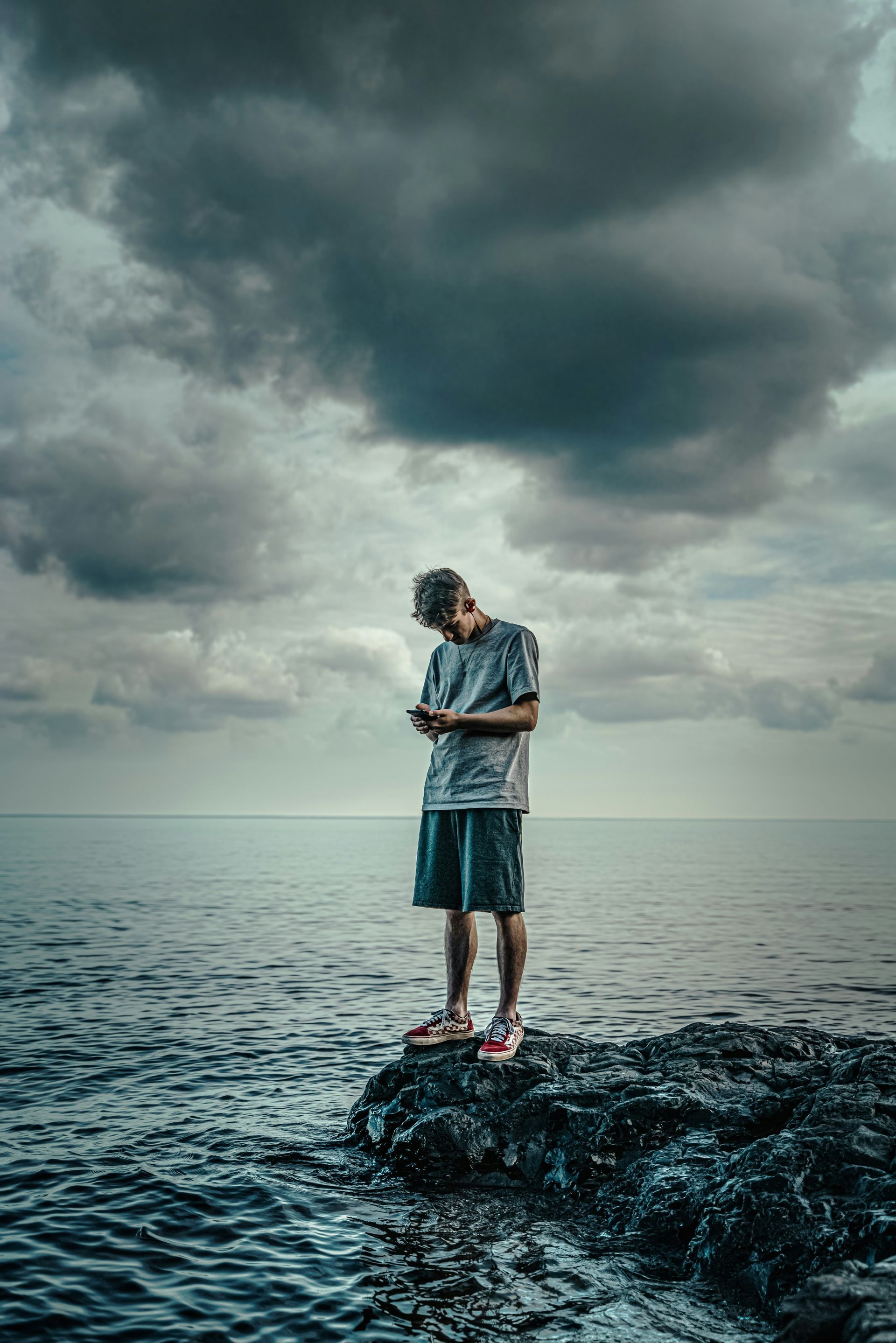 Person standing on rocky shore under dark storm clouds by calm water