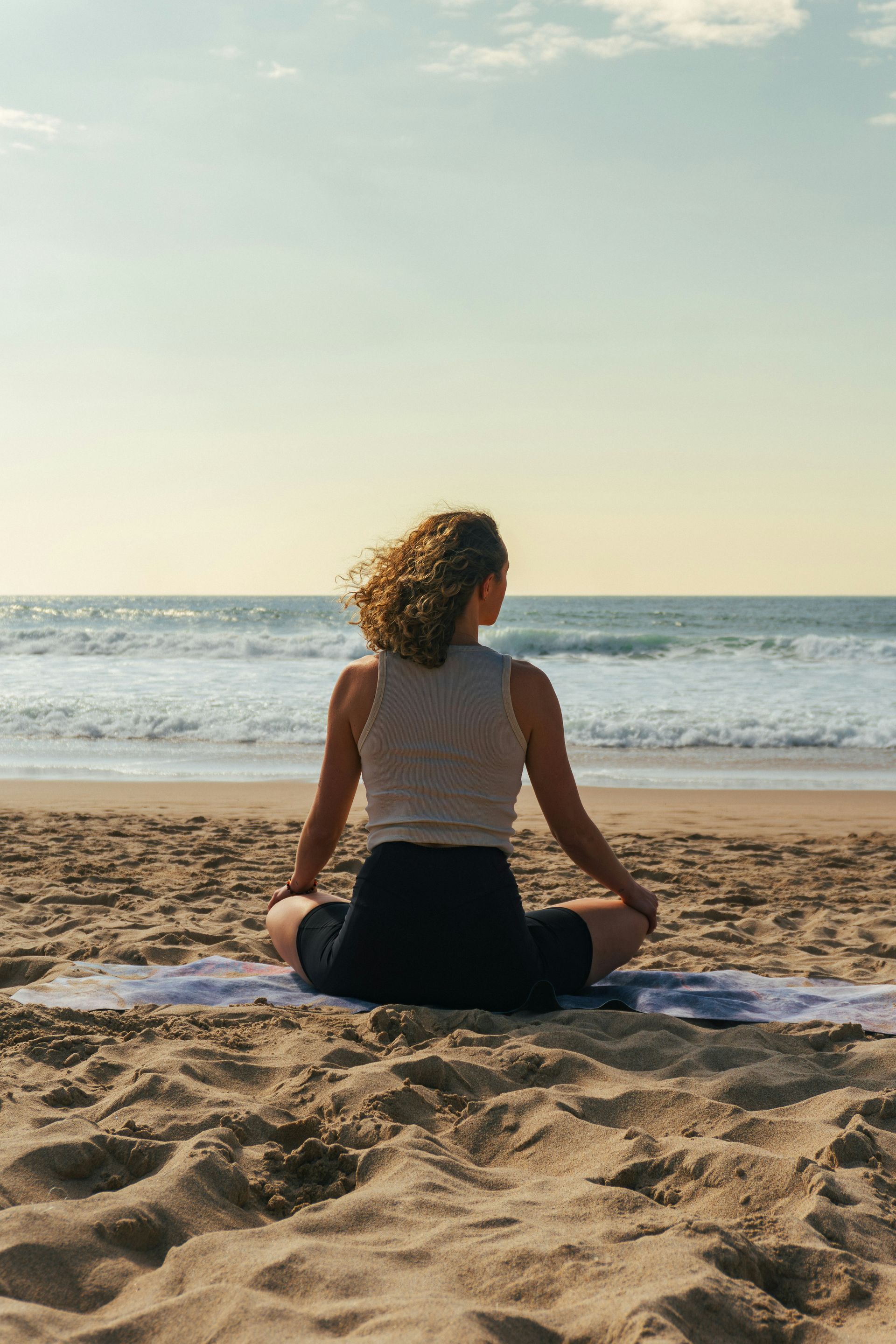 Person meditating on a beach at sunrise, facing the ocean, with a towel beside them