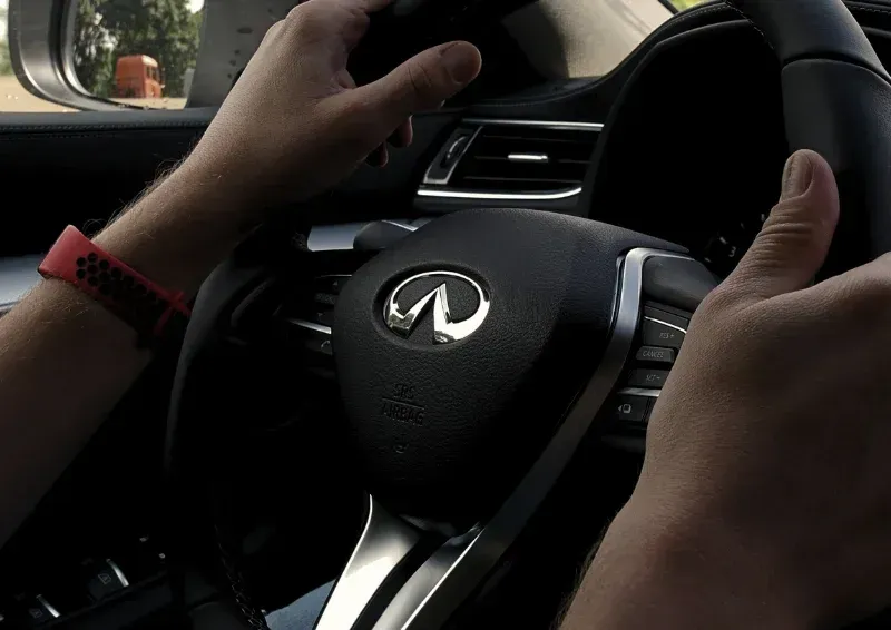 Hands on a black Infiniti steering wheel inside a car, with a red wristband visible.