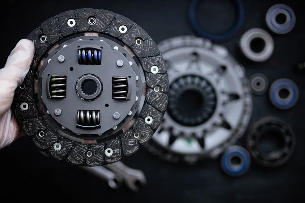 Hand holding a car clutch disc, with a larger clutch assembly and bearings in the background