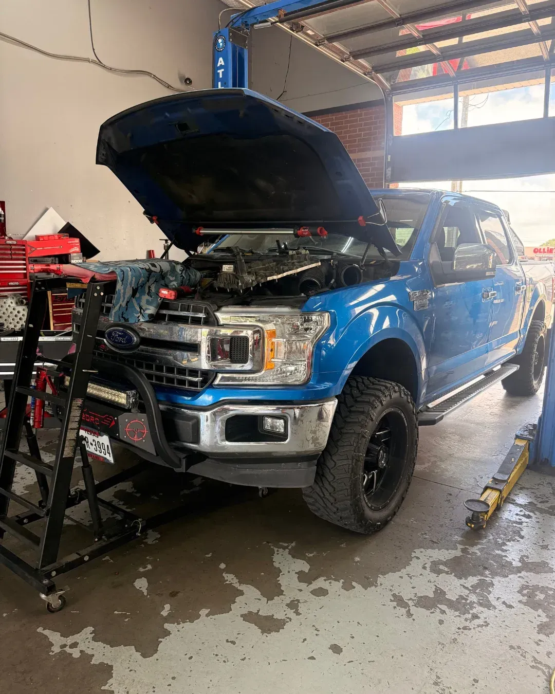 Four mechanics standing in an auto shop beside a silver car with its hood open and a tire machine nearby