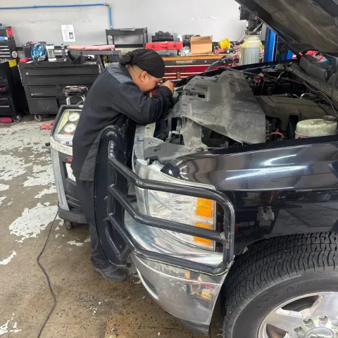 Mechanic servicing a car engine in a workshop, with gauges and blue hoses attached to the open hood.