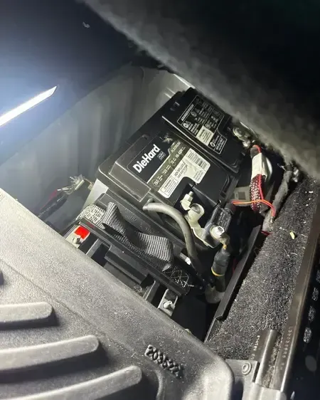 Close-up of a car battery in a dusty engine compartment with cables and wiring visible.