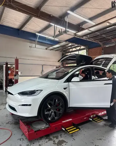 White Tesla on a red lift in an auto shop, with its hood and trunk open and a person inspecting inside.