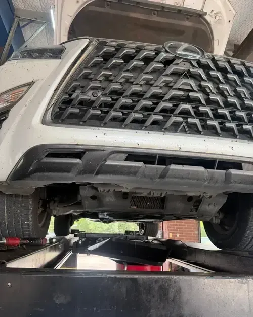 White SUV front end raised on a lift, showing the grille and underside in a repair shop