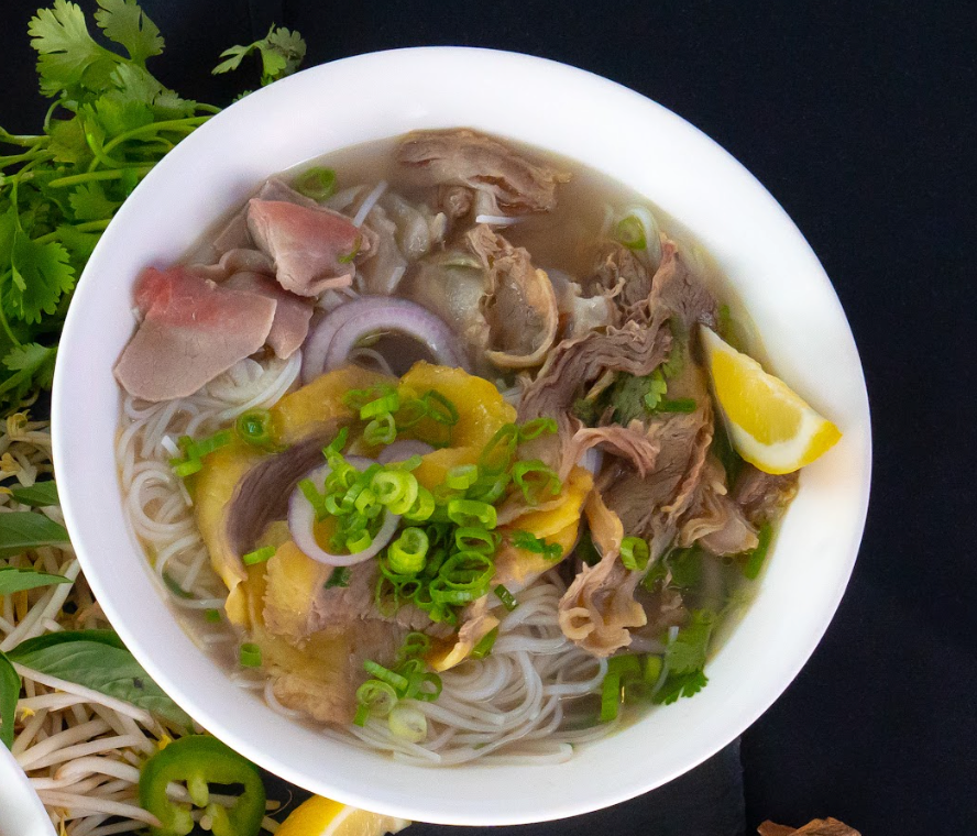 Bowl of noodle soup topped with sliced meat, herbs, and lemon wedges on a black surface