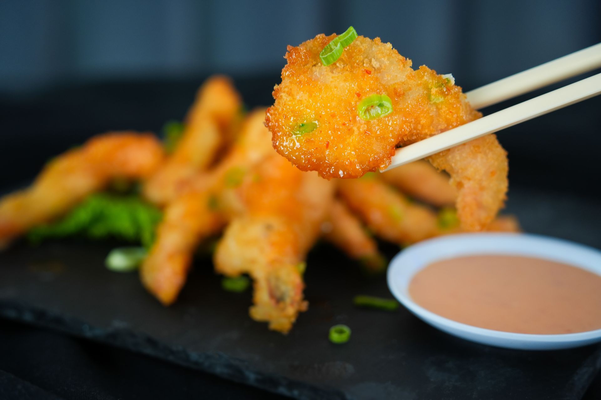 Chopsticks lifting a crispy orange fried appetizer with dipping sauce on a black plate