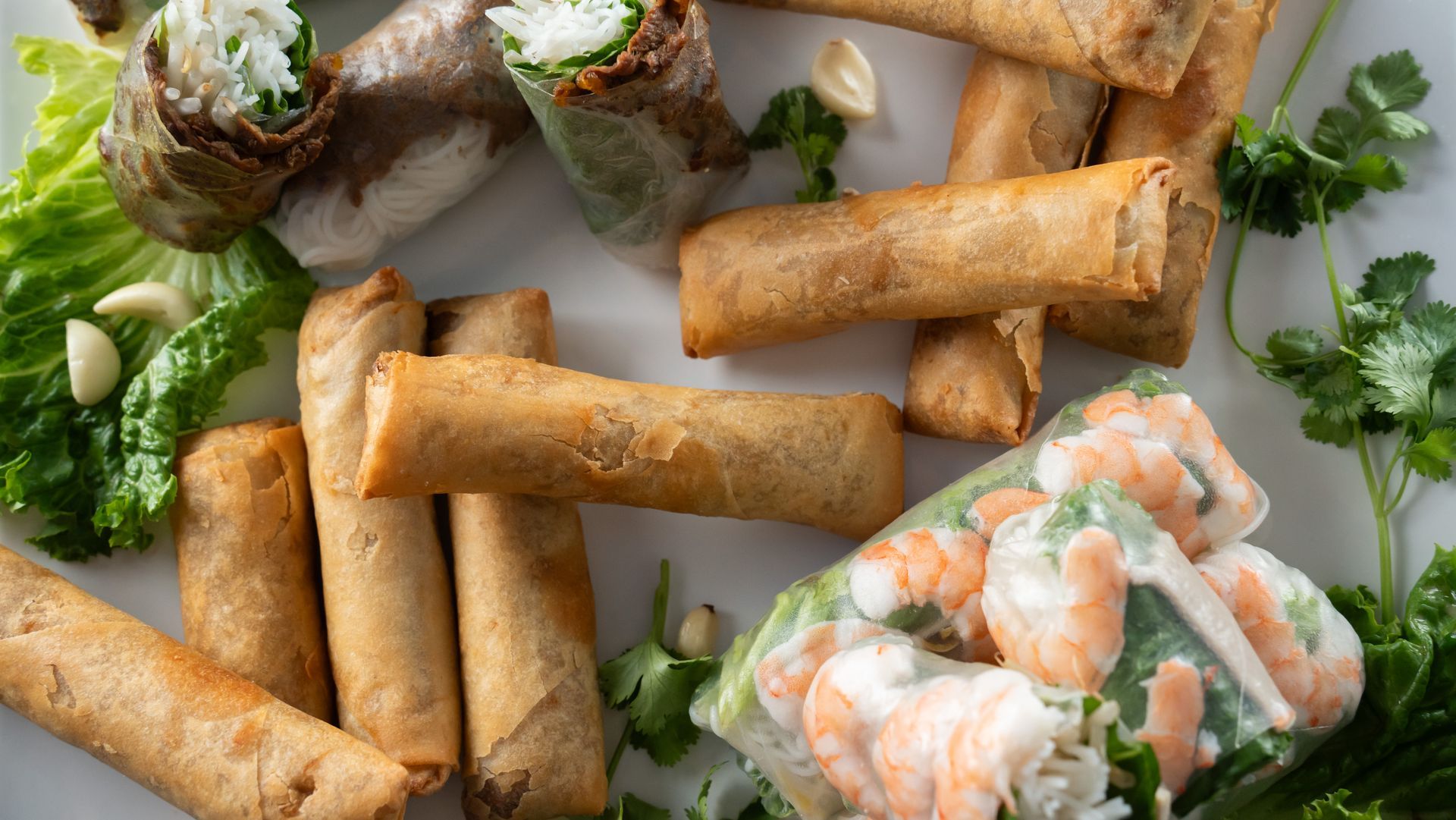 Assorted spring rolls on parchment with shrimp rolls, herbs, and dipping sauce on a light table