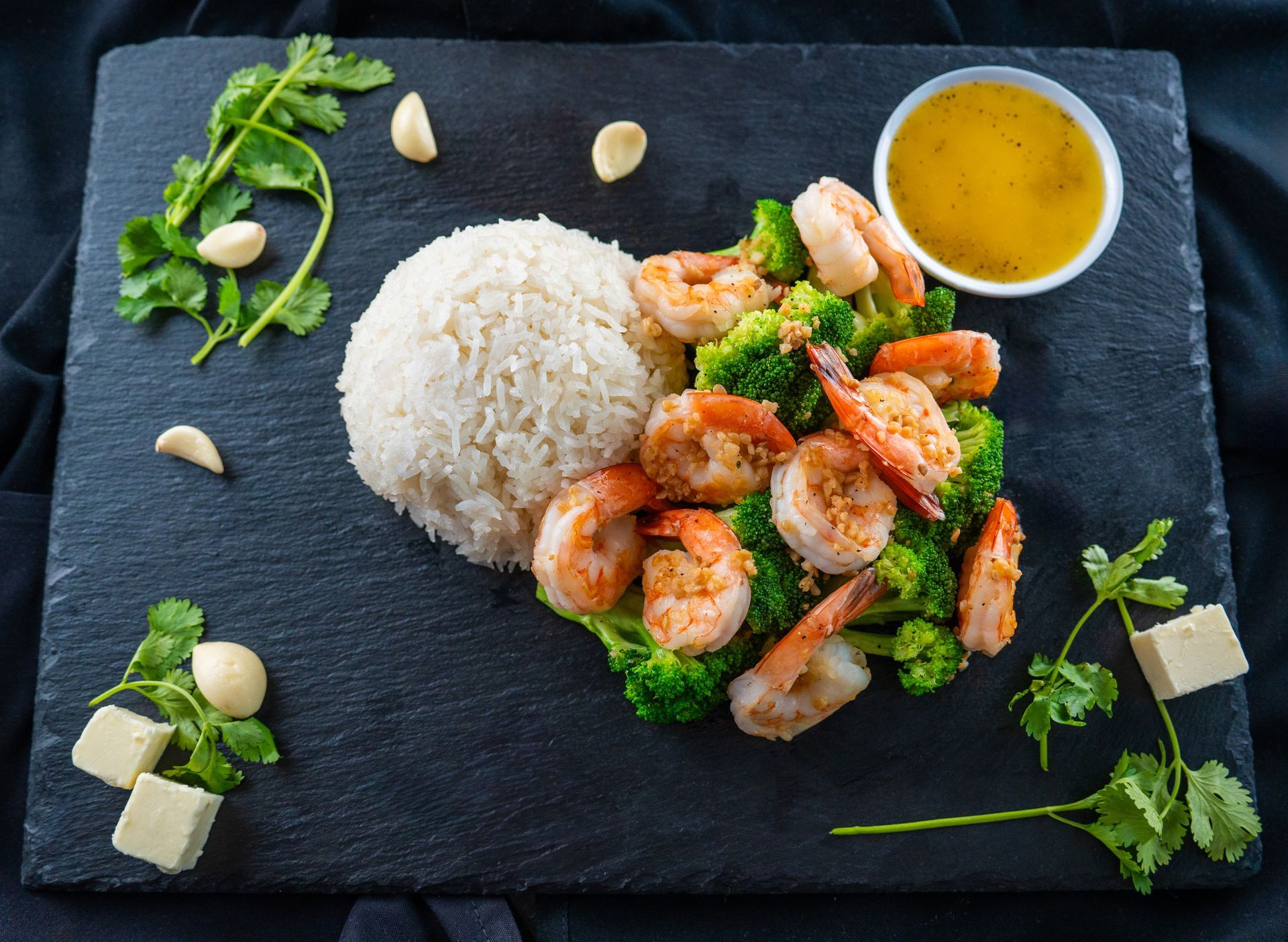 Rice with shrimp and broccoli, garnished with herbs, alongside a small bowl of sauce on a black plate.