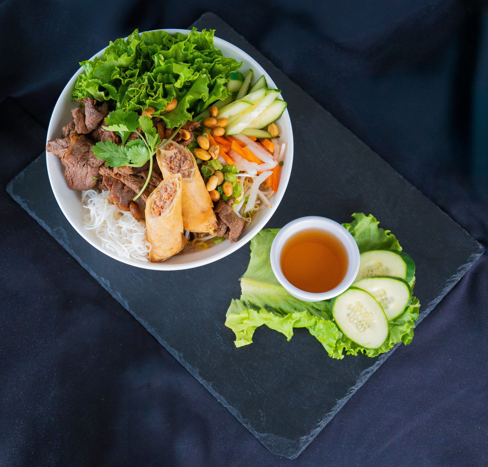 Vietnamese noodle bowl with beef, herbs, vegetables, and dipping sauce on a dark table