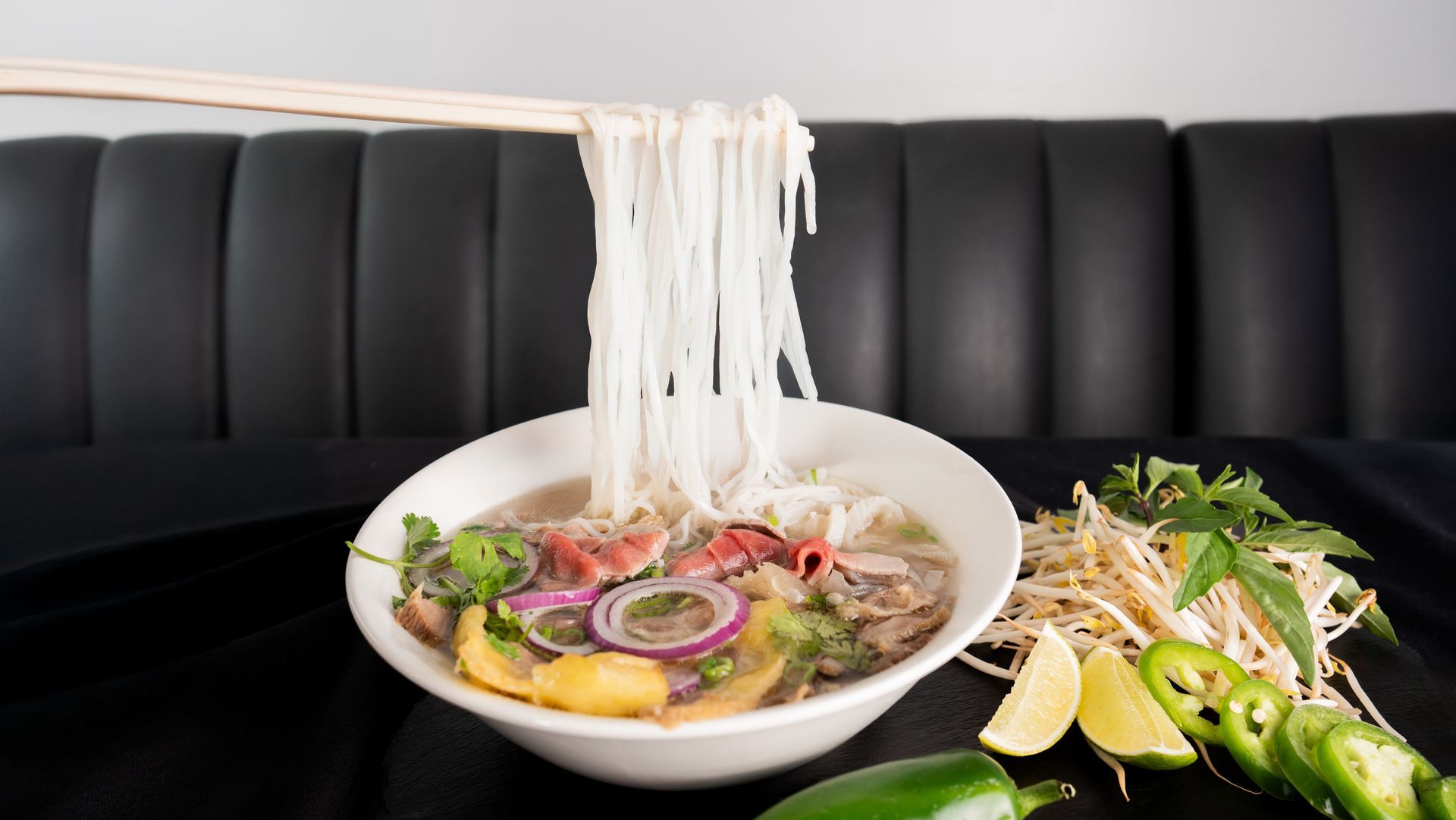 Bowl of noodle soup with sliced onions and herbs on a black table beside lime and chili garnish
