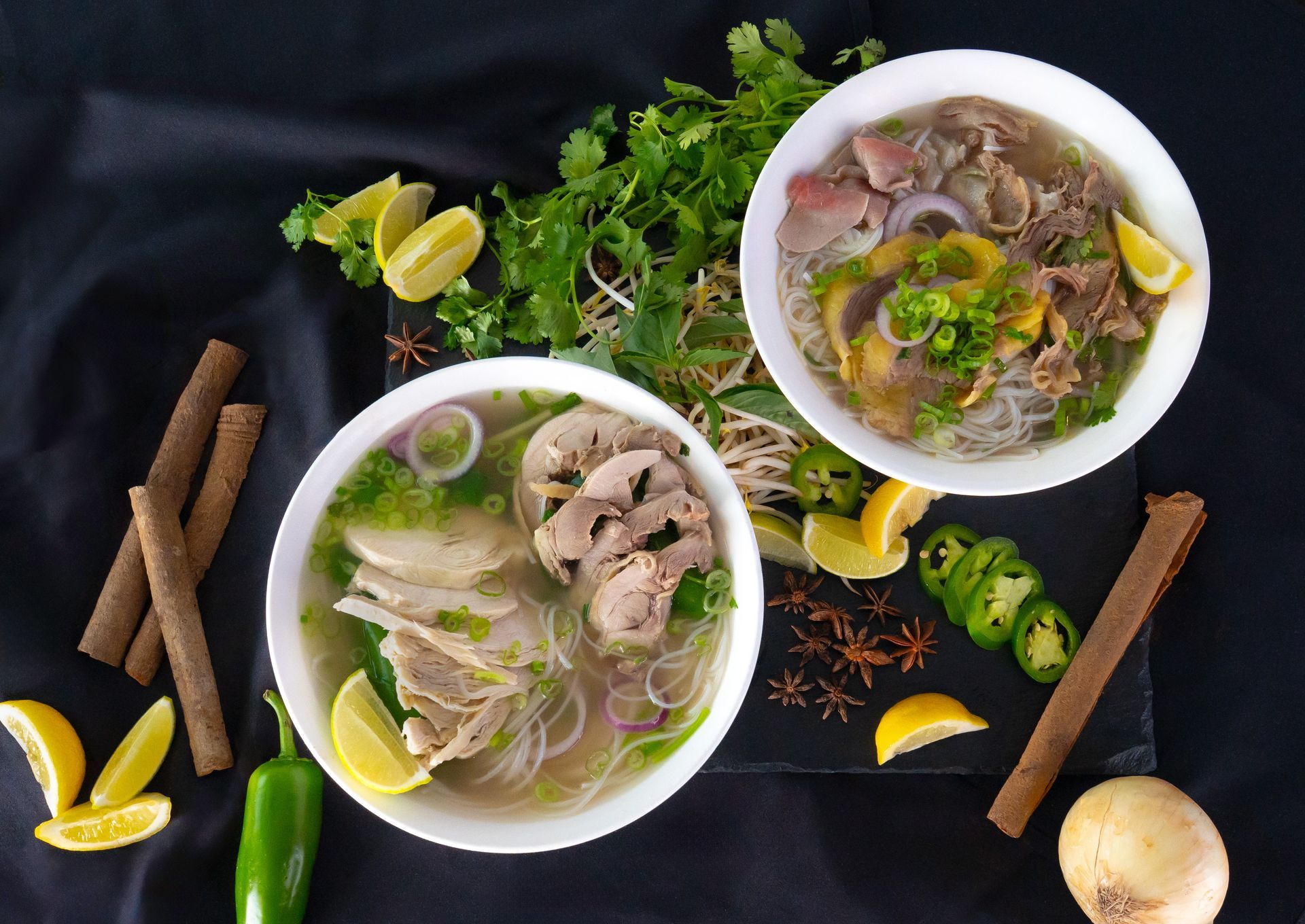 Two bowls of pho with lime and herbs on a dark table, surrounded by chilies, cinnamon sticks, and spices