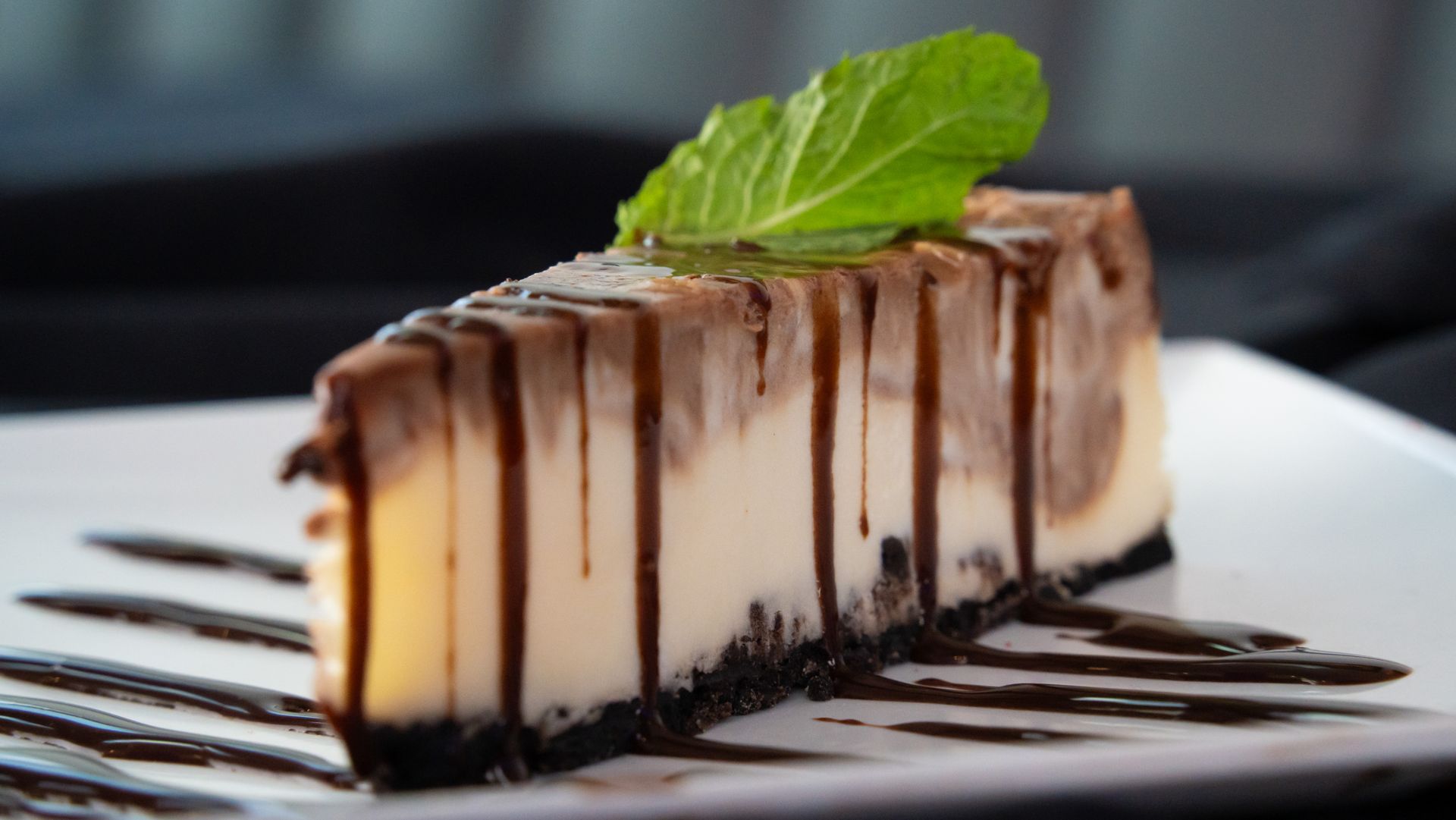 Slice of cheesecake drizzled with chocolate sauce on a white plate, topped with a mint leaf