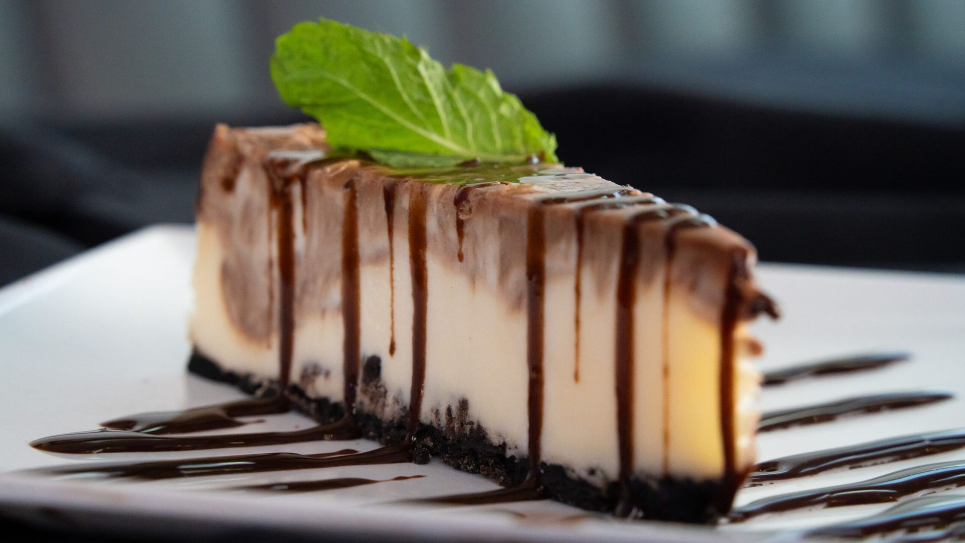 Slice of cheesecake drizzled with chocolate sauce, topped with mint on a white plate