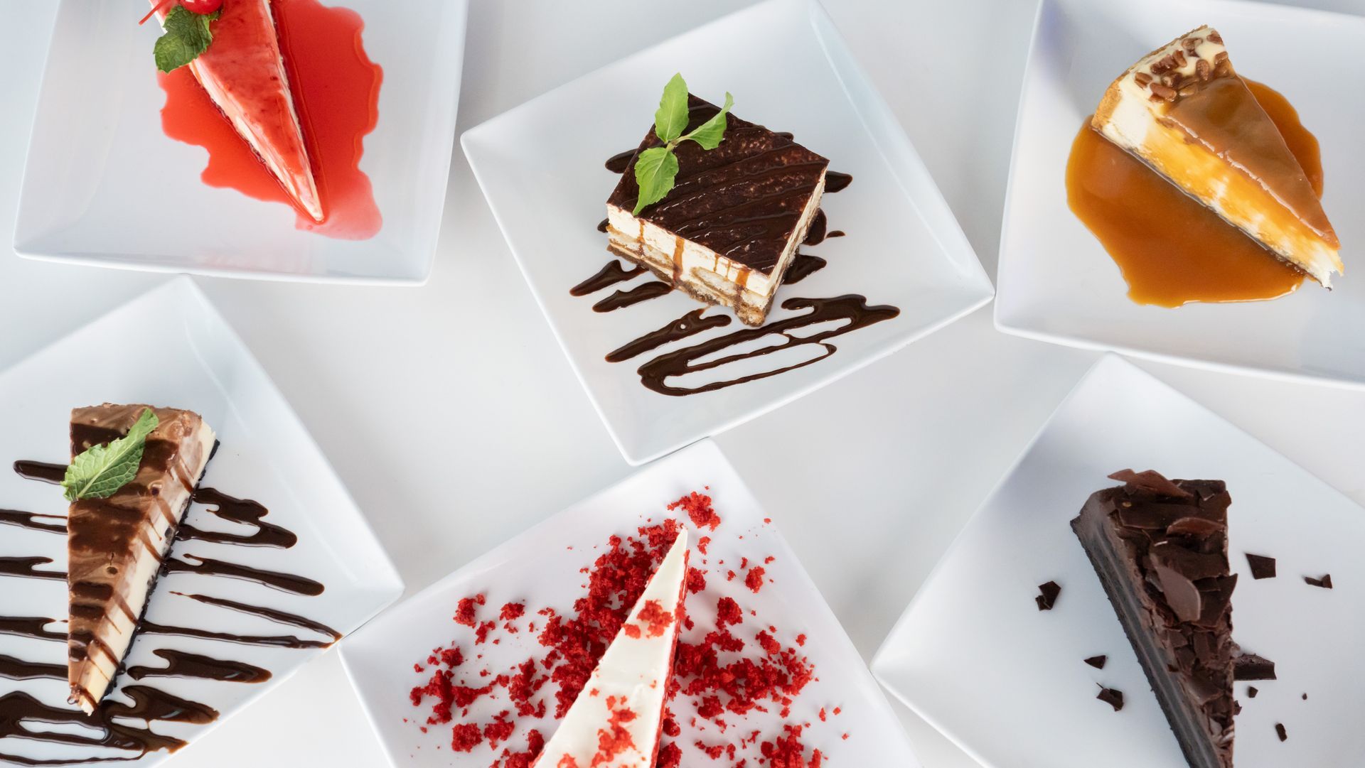 Assorted sliced desserts on white plates, including chocolate, caramel, strawberry, and cherry-topped cakes.