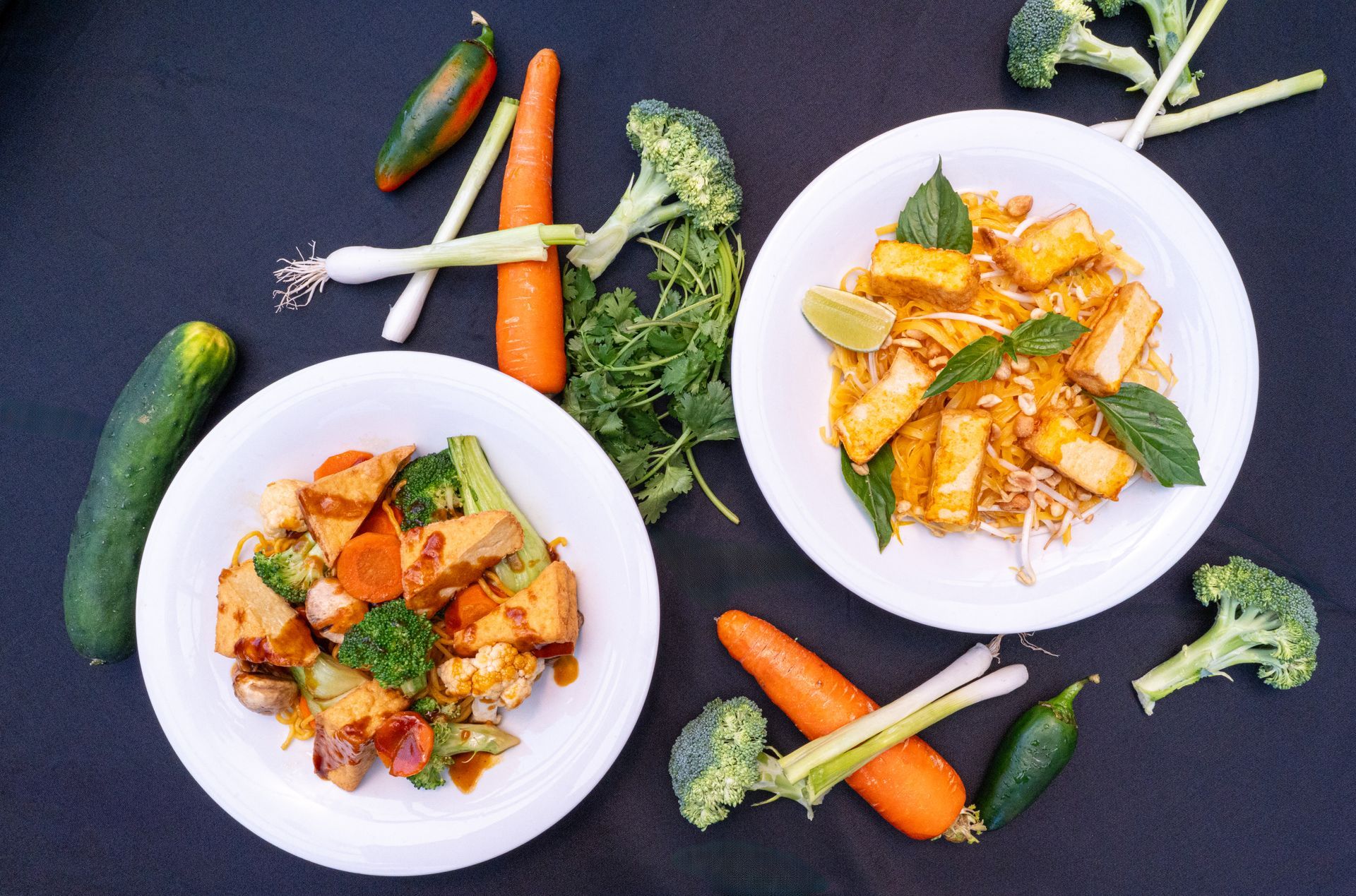 Two plates of pasta with basil, surrounded by cucumbers, carrots, and broccoli on a dark background