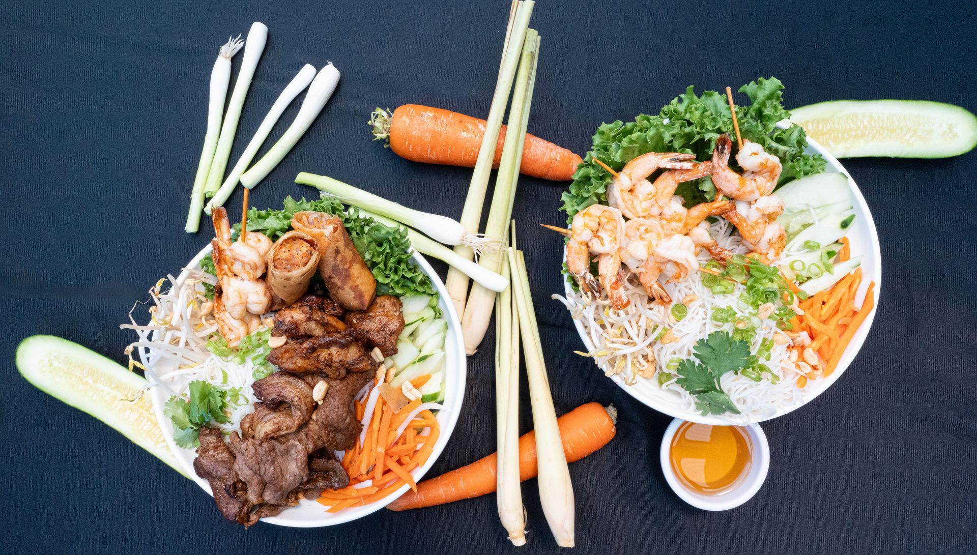 Two bowls of noodle salad with meat and shrimp, chopsticks, carrots, and cucumber on a dark table.