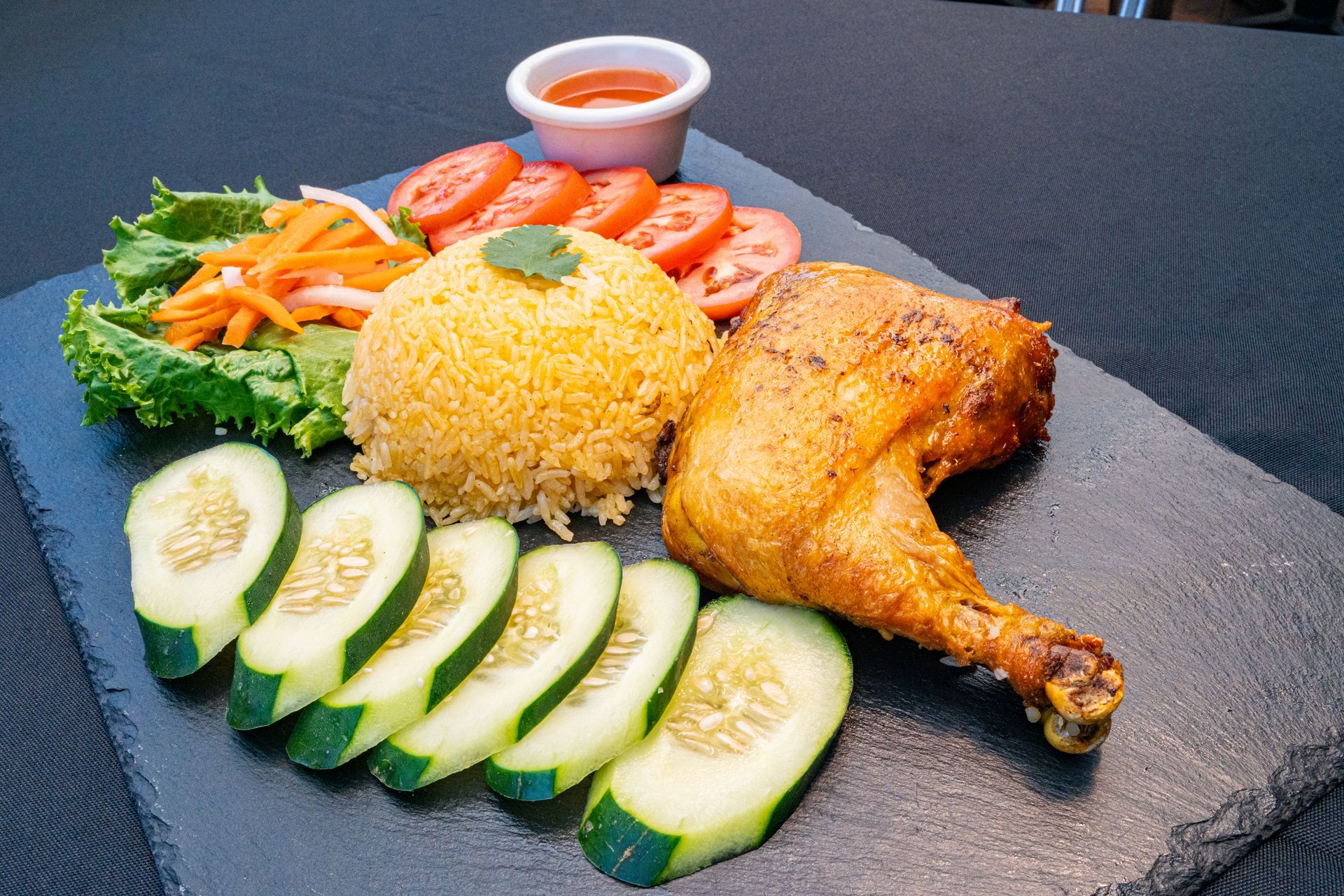 Grilled chicken leg with rice, cucumber slices, tomatoes, carrots, and dipping sauce on a dark slate plate