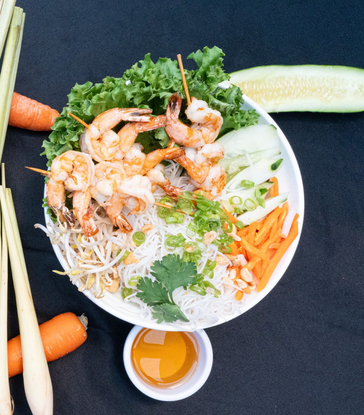 Shrimp spring roll bowl with lettuce, herbs, carrots, noodles, and dipping sauce on dark background