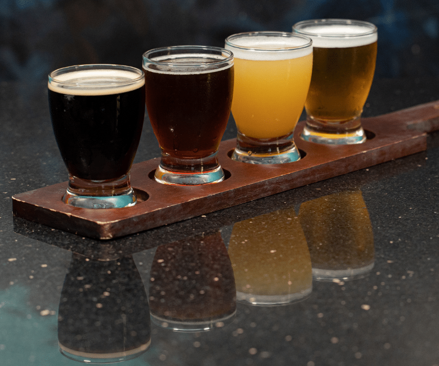 Four small beer samples in glasses on a wooden flight tray on a dark countertop.